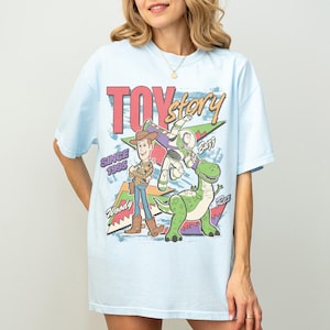 Op de afbeelding: Lichtblauw T-shirt met de tekst "Toy Story" in rood en groen, met Woody, Buzz Lightyear en Rex de dinosaurus. De tekst "Since 1995" is ook zichtbaar.