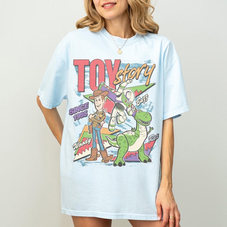 Op de afbeelding: Lichtblauw T-shirt met de tekst "Toy Story" in rood en groen, met Woody, Buzz Lightyear en Rex de dinosaurus. De tekst "Since 1995" is ook zichtbaar.