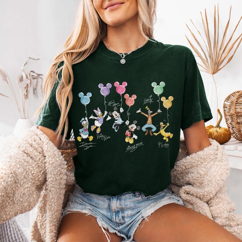 Pode incluir: T-shirt verde escuro com um gr&aacute;fico colorido de personagens da Disney segurando bal&otilde;es em forma de Mickey Mouse. Os personagens incluem Pato Donald, Margarida, Minnie, Mickey Mouse, Pateta e Pluto. Os nomes dos personagens est&atilde;o escritos abaixo.