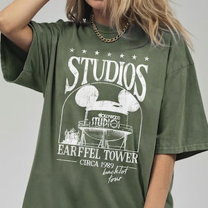 May include: Olive green t-shirt with a white graphic featuring the word "STUDIOS" above a Mickey Mouse-shaped water tower design. The graphic includes the text "HOLLYWOOD STUDIO!" and "EARFFEL TOWER CIRCA 1989 backlot tour."