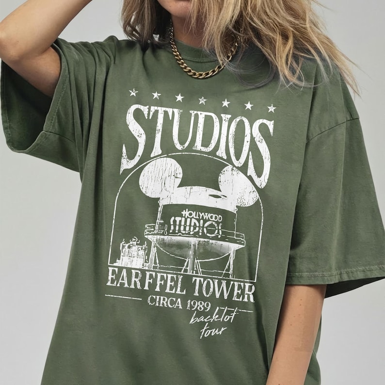 May include: Olive green t-shirt with a white graphic featuring the word "STUDIOS" above a Mickey Mouse-shaped water tower design. The graphic includes the text "HOLLYWOOD STUDIO!" and "EARFFEL TOWER CIRCA 1989 backlot tour."