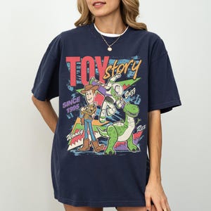 Op de afbeelding: Marineblauwe t-shirt met een kleurrijke afbeelding van Woody, Buzz Lightyear en Rex uit Toy Story. De tekst "TOY STORY" en "SINCE 1995" zijn ook afgebeeld. De t-shirt heeft een ronde hals en korte mouwen.