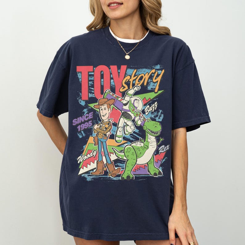 Op de afbeelding: Marineblauwe t-shirt met een kleurrijke afbeelding van Woody, Buzz Lightyear en Rex uit Toy Story. De tekst "TOY STORY" en "SINCE 1995" zijn ook afgebeeld. De t-shirt heeft een ronde hals en korte mouwen.
