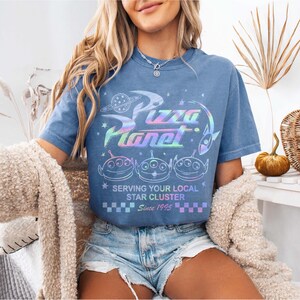 May include: A blue t-shirt featuring the Pizza Planet logo in a holographic design. Below the logo are three alien characters and the text "Serving Your Local Star Cluster Since 1995". The shirt has short sleeves.