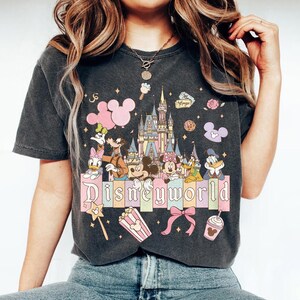 Puede incluir: Camiseta gris oscuro con un dise&ntilde;o colorido de Disney World. El gr&aacute;fico incluye personajes de Disney, un castillo y el texto "Disneyworld" en colores pastel. Elementos adicionales: un lazo rosa, palomitas de ma&iacute;z y una varita m&aacute;gica.