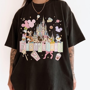 May include: Black t-shirt featuring a colorful Disneyland graphic. The design includes a castle, Mickey Mouse, Minnie Mouse, Donald Duck, and other Disney characters. The word "Disneyland" is displayed in pastel colors.