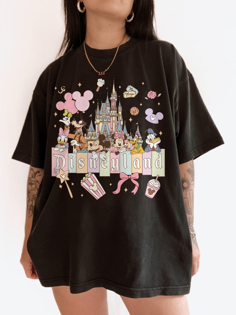 May include: Black t-shirt featuring a colorful Disneyland graphic. The design includes a castle, Mickey Mouse, Minnie Mouse, Donald Duck, and other Disney characters. The word "Disneyland" is displayed in pastel colors.
