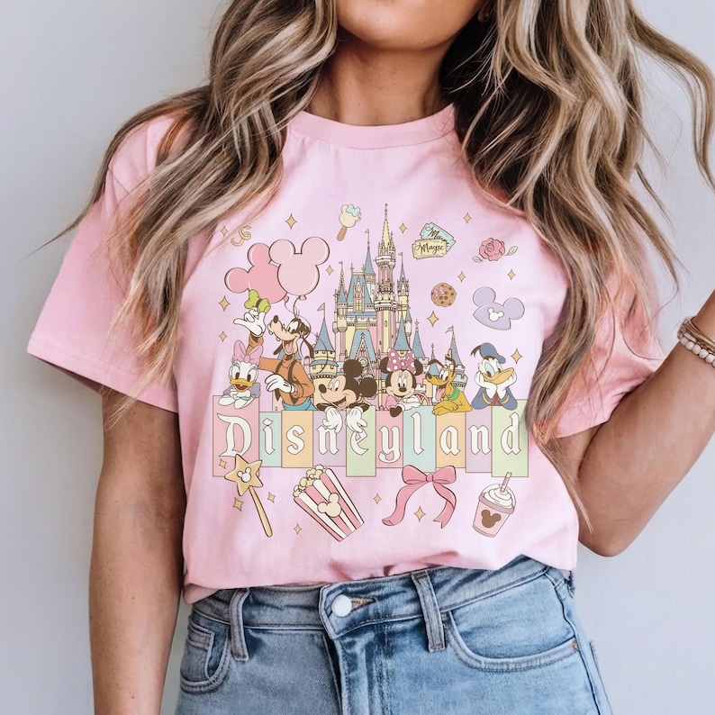 May include: Pink t-shirt featuring a Disneyland graphic. The design includes a castle, Mickey Mouse, Minnie Mouse, Donald Duck, Goofy, and other characters. The word "Disneyland" is written across the bottom in a pastel font. Other graphics include a bow, popcorn, and a magic wand.