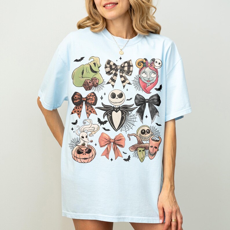 May include: A light blue t-shirt featuring a graphic print of characters and bows from The Nightmare Before Christmas. The design includes Jack Skellington, Sally, Zero, Oogie Boogie, and Lock, Shock, and Barrel, interspersed with black bats and spiderwebs.