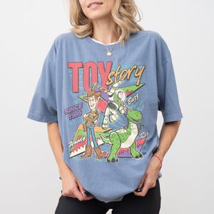 Op de afbeelding: Blauw T-shirt met Toy Story-logo en de personages Woody, Buzz en Rex. De afbeelding bevat de tekst "Toy Story Since 1995". Het shirt heeft korte mouwen en een losse pasvorm.