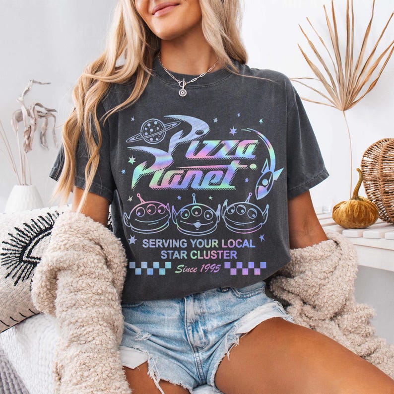 May include: Dark gray t-shirt with a holographic "Pizza Planet" graphic, featuring alien characters and the text "Serving Your Local Star Cluster Since 1995." The shirt has a crew neck and short sleeves.