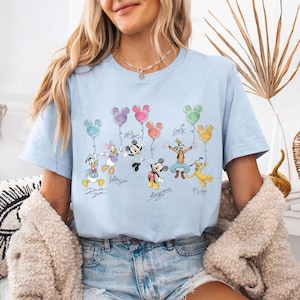 Pode incluir: Camiseta azul clara com um desenho de personagens da Disney segurando bal&otilde;es em forma de Mickey Mouse. O desenho inclui Pato Donald, Margarida, Minnie, Mickey, Pateta e Pluto. Os nomes est&atilde;o escritos abaixo.