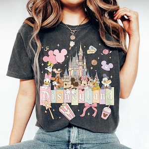 May include: Dark gray t-shirt featuring a colorful Disneyland graphic. The design includes Disney characters, a castle, and the word "Disneyland" in pastel colors. Additional graphics include a bow, popcorn, and a drink.