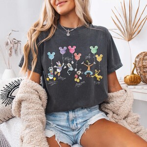 Pode incluir: Camiseta cinza escuro com personagens da Disney segurando bal&otilde;es coloridos. O design inclui Pato Donald, Margarida, Minnie Mouse, Mickey Mouse, Pateta e Pluto. Cada personagem tem sua assinatura abaixo.