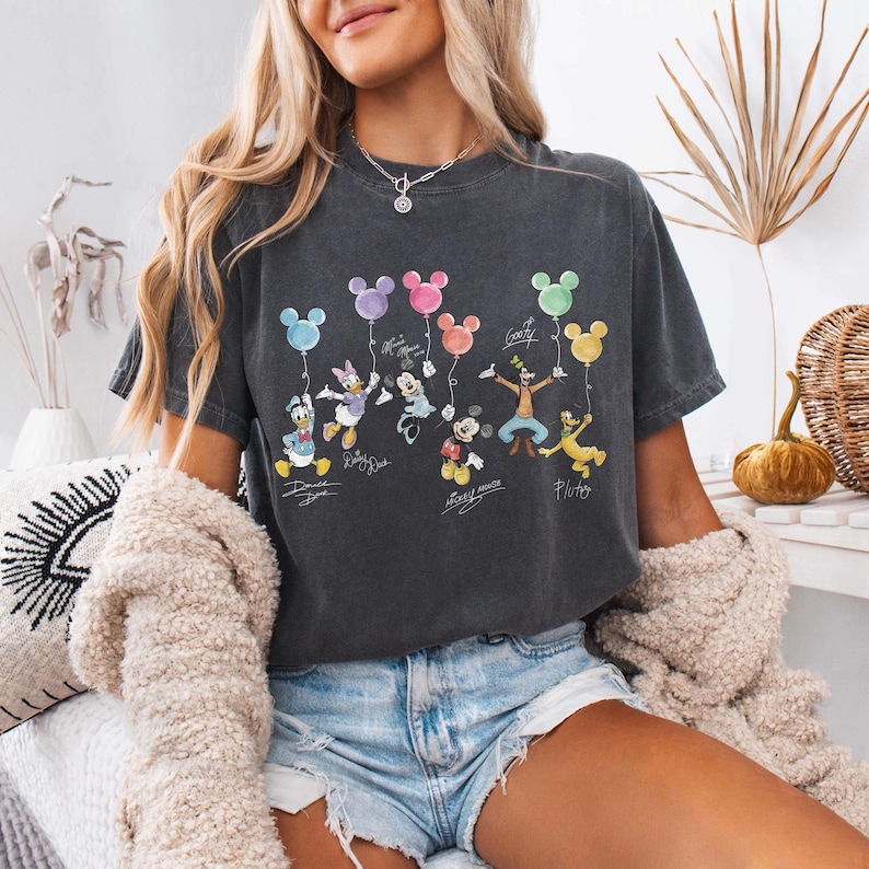 Pode incluir: Camiseta cinza escuro com personagens da Disney segurando bal&otilde;es coloridos. O design inclui Pato Donald, Margarida, Minnie Mouse, Mickey Mouse, Pateta e Pluto. Cada personagem tem sua assinatura abaixo.