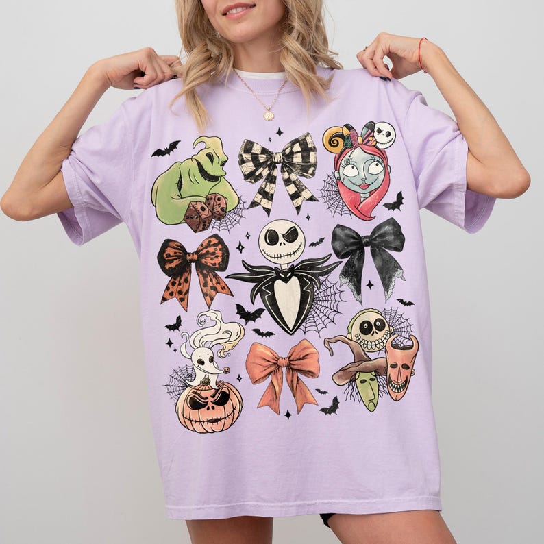 May include: A light purple t-shirt featuring a graphic print of characters and bows from The Nightmare Before Christmas. The design includes Jack Skellington, Sally, Oogie Boogie, Zero, and various bows, all surrounded by small bats and spiderwebs.