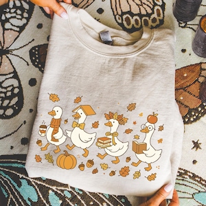 May include: Beige sweatshirt featuring a whimsical autumn design. The graphic depicts four cartoon geese, each adorned with fall-themed accessories like scarves, books, and an apple. Scattered leaves and a pumpkin complete the seasonal illustration.