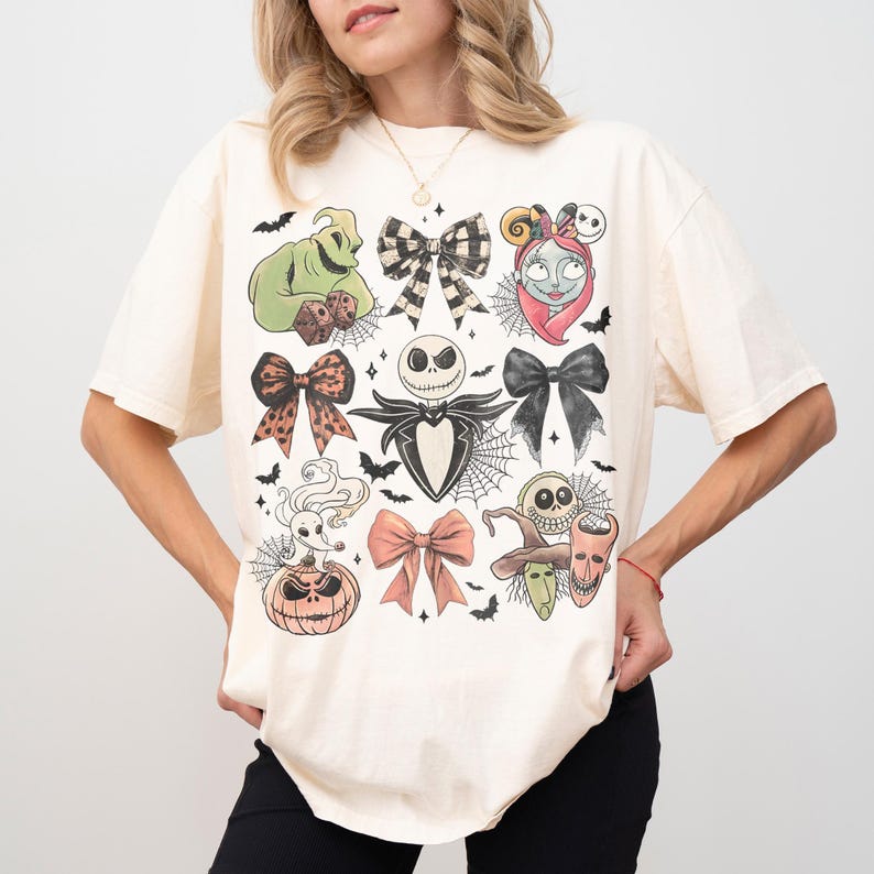 May include: A cream-colored short-sleeve t-shirt featuring a graphic print of various characters and bows. The design includes a green sack character, a skull-faced figure, a stitched doll, a ghost dog on a pumpkin, and several decorative bows.