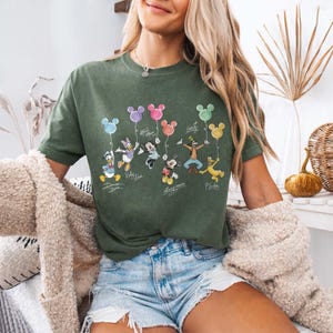 Pode incluir: Camiseta verde-oliva com personagens da Disney segurando bal&otilde;es em forma de Mickey Mouse. Os personagens incluem Pato Donald, Margarida, Mickey Mouse, Minnie, Pateta e Pluto. O nome de cada personagem est&aacute; escrito abaixo.