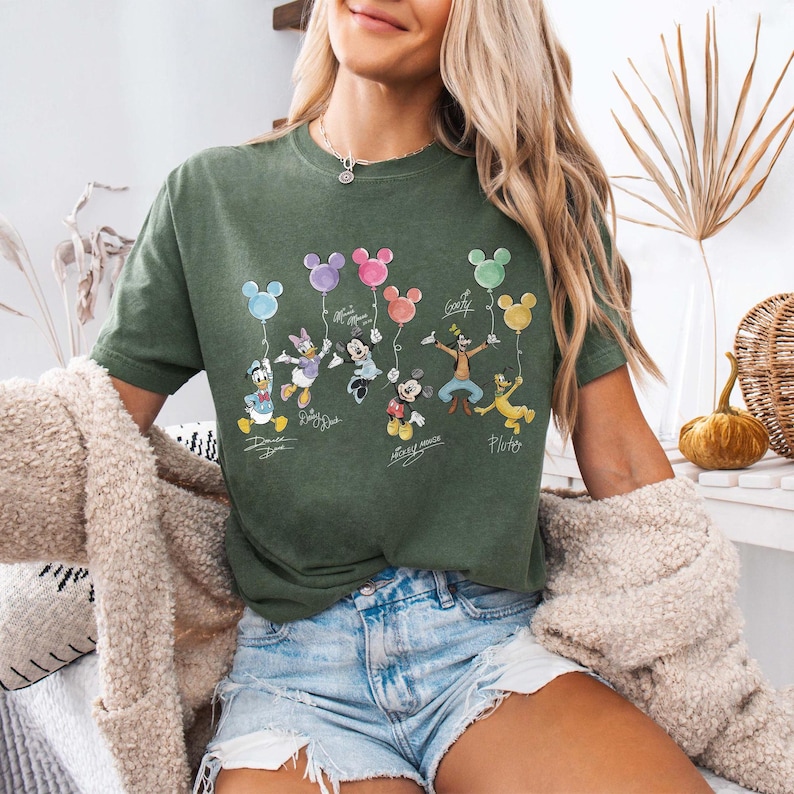 Pode incluir: Camiseta verde-oliva com personagens da Disney segurando bal&otilde;es em forma de Mickey Mouse. Os personagens incluem Pato Donald, Margarida, Mickey Mouse, Minnie, Pateta e Pluto. O nome de cada personagem est&aacute; escrito abaixo.