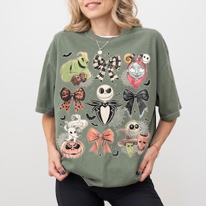 May include: An olive green short-sleeve t-shirt featuring a graphic print of characters and bows from The Nightmare Before Christmas. The design includes Jack Skellington, Sally, Oogie Boogie, Zero, and various bows arranged in a grid pattern.