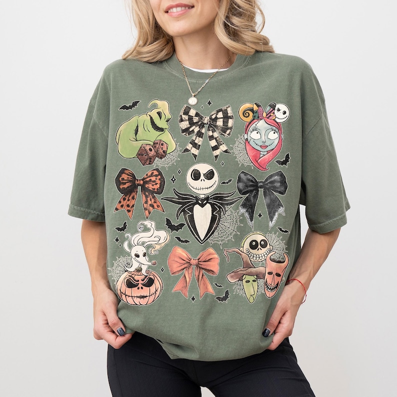 May include: An olive green short-sleeve t-shirt featuring a graphic print of characters and bows from The Nightmare Before Christmas. The design includes Jack Skellington, Sally, Oogie Boogie, Zero, and various bows arranged in a grid pattern.
