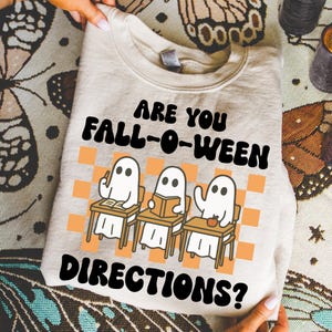 Puede incluir: Una sudadera beige con el texto "ARE YOU FALL-O-WEEN DIRECTIONS?" en negro. El diseño presenta tres fantasmas de dibujos animados en escritorios frente a un fondo a cuadros naranja y blanco. Los fantasmas están leyendo y escribiendo.