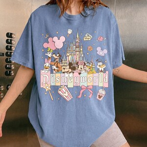 Puede incluir: Camiseta azul claro con un dise&ntilde;o de "Disneyworld". El dise&ntilde;o presenta personajes de Disney como Mickey y Minnie Mouse, Donald Duck y Goofy, junto con un castillo y el texto "Disneyworld". Se utilizan colores pastel.