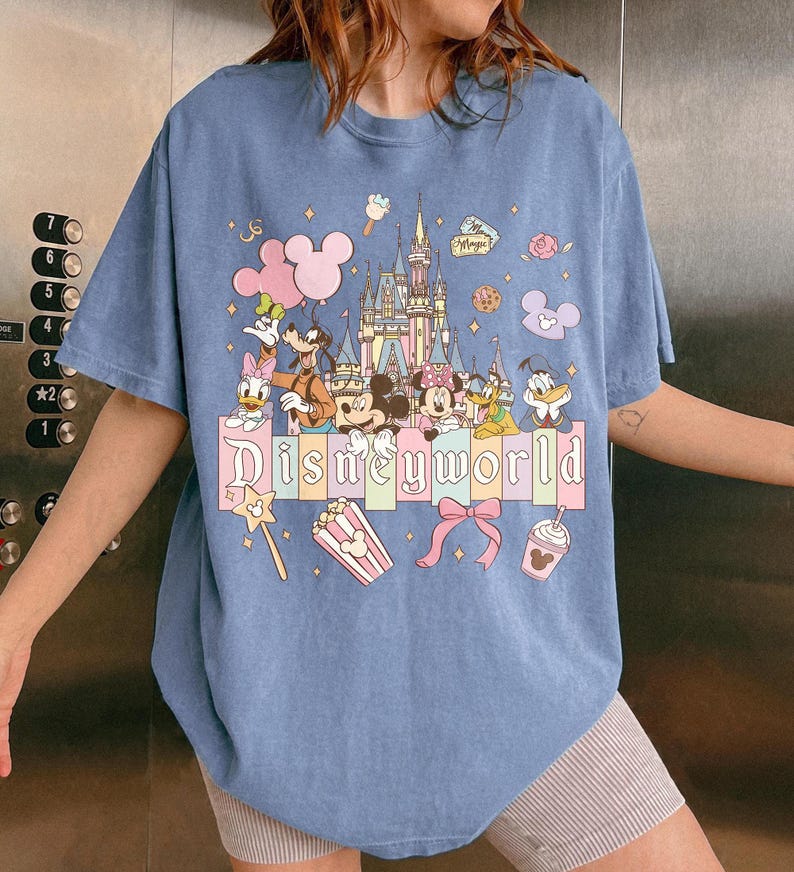Puede incluir: Camiseta azul claro con un dise&ntilde;o de "Disneyworld". El dise&ntilde;o presenta personajes de Disney como Mickey y Minnie Mouse, Donald Duck y Goofy, junto con un castillo y el texto "Disneyworld". Se utilizan colores pastel.