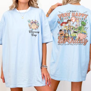 May include: Light blue t-shirt with a graphic featuring a cartoon character and the text "Birthday Boy" on the front. The back of the shirt has a colorful design with the text "Jackson's Very Happy 7th Birthday" and cartoon characters.
