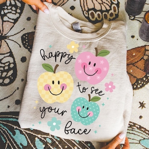 Op de afbeelding: Een beige sweatshirt met de tekst "happy to see your face" en drie lachende appelontwerpen in geel, roze en blauw. De appels hebben groene bladeren en zijn versierd met bloemen en sterren. De sweatshirt is opgevouwen.