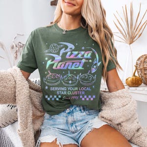 May include: Green t-shirt with a holographic "Pizza Planet" graphic, featuring alien characters and the text "Serving Your Local Star Cluster Since 1995." The shirt has a vintage, worn look.