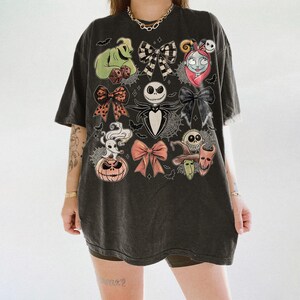 May include: A charcoal gray oversized t-shirt featuring a graphic print of characters from The Nightmare Before Christmas, including Jack Skellington, Sally, Oogie Boogie, and Zero, interspersed with various patterned bows and small bats.