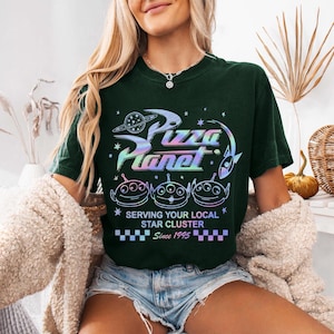 May include: Dark green t-shirt featuring the text "Pizza Planet" in a holographic design, along with three alien characters. The shirt also includes the text "Serving Your Local Star Cluster Since 1995".