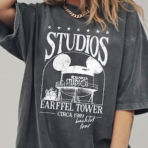 May include: Dark gray t-shirt with a distressed graphic featuring the text "STUDIOS" and "EARFFEL TOWER CIRCA 1989 backlot tour". The graphic includes a stylized water tower design with Mickey Mouse ears. The shirt has short sleeves.