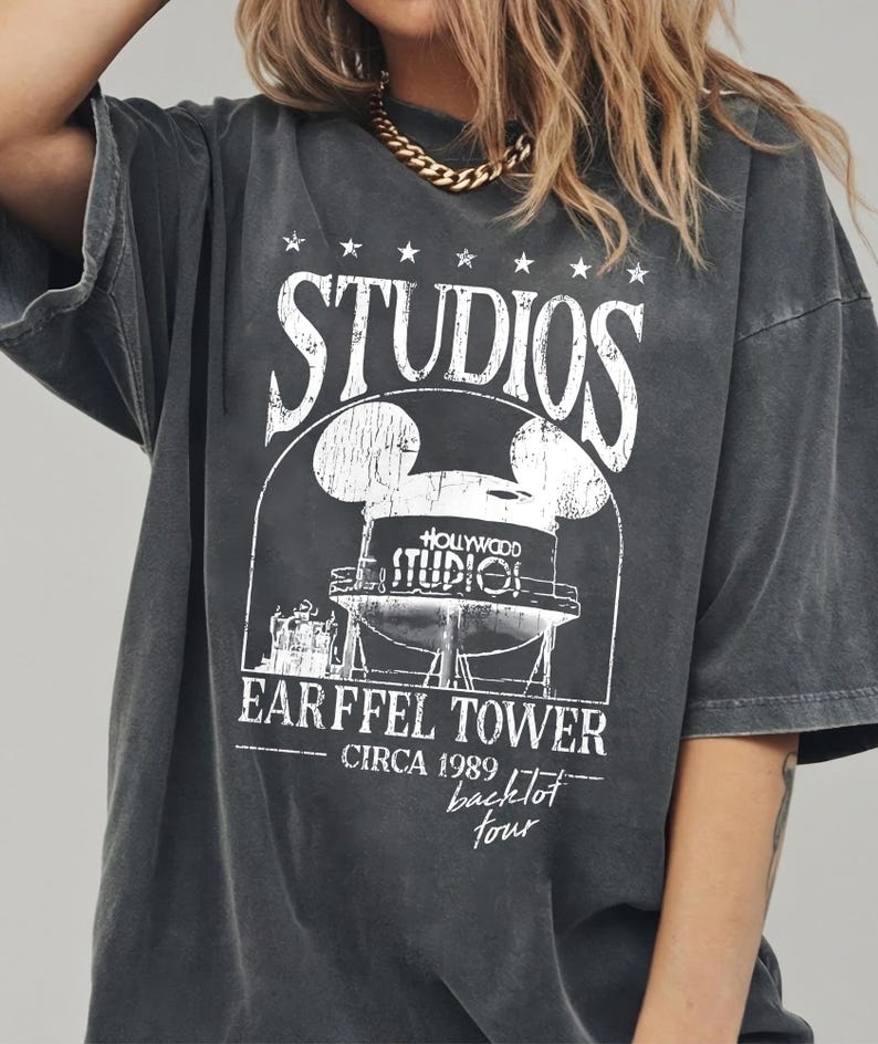 May include: Dark gray t-shirt with a distressed graphic featuring the text "STUDIOS" and "EARFFEL TOWER CIRCA 1989 backlot tour". The graphic includes a stylized water tower design with Mickey Mouse ears. The shirt has short sleeves.