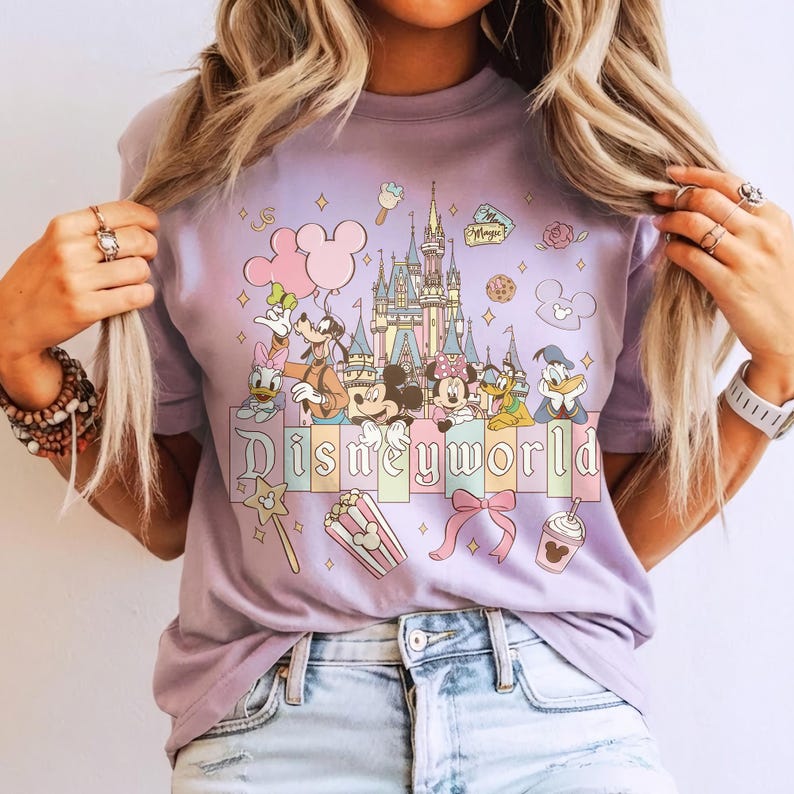 Puede incluir: Una camiseta de color lila con un gr&aacute;fico colorido de personajes de Disney, incluyendo Mickey y Minnie Mouse, Donald y Daisy Duck y Goofy. La palabra "Disneyworld" est&aacute; impresa en la parte inferior, con un castillo y otros iconos.