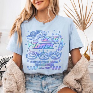 May include: Light blue t-shirt featuring a holographic Pizza Planet logo with the text "Serving Your Local Star Cluster Since 1995". The design includes three alien characters and star graphics.