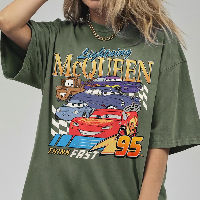 Puede incluir: Camiseta verde oliva con un gr&aacute;fico de Rayo McQueen y otros personajes de Cars. El dise&ntilde;o incluye el texto "Lightning McQueen" y "Think Fast 95". La camiseta tiene un aspecto vintage y ligeramente descolorido.
