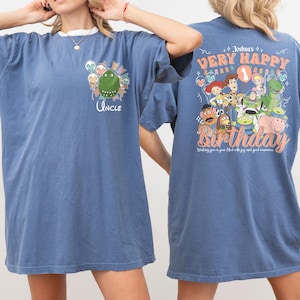 May include: Blue t-shirt with a white collar featuring a cartoon dinosaur and the word "Uncle". The back of the shirt has a graphic with the text "Very Happy Birthday" and various cartoon characters.