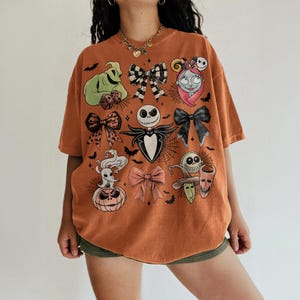 May include: An orange short-sleeve t-shirt featuring a graphic print of various characters and bows. The design includes a green figure, a skull-faced character, a ghost dog, a pumpkin, and several decorative bows arranged across the front.