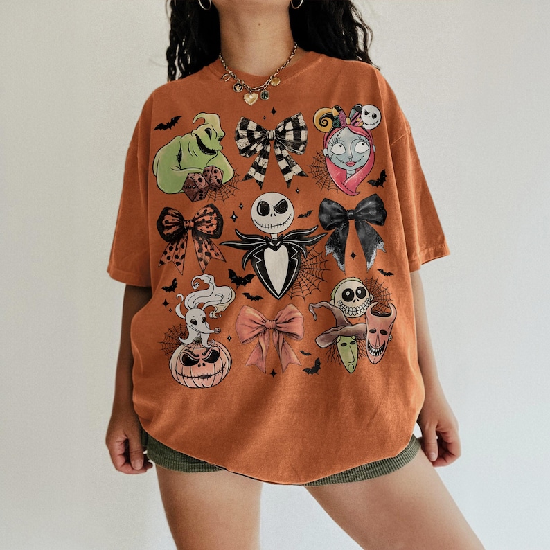 May include: An orange short-sleeve t-shirt featuring a graphic print of various characters and bows. The design includes a green figure, a skull-faced character, a ghost dog, a pumpkin, and several decorative bows arranged across the front.