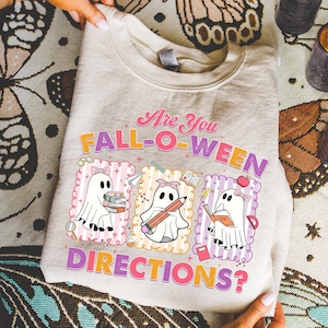 May include: A light beige sweatshirt with the text "Are You FALL-O-WEEN DIRECTIONS?" in pink, orange, and purple. The design features three cartoon ghosts with school-themed accessories, including books, pencils, and an apple.