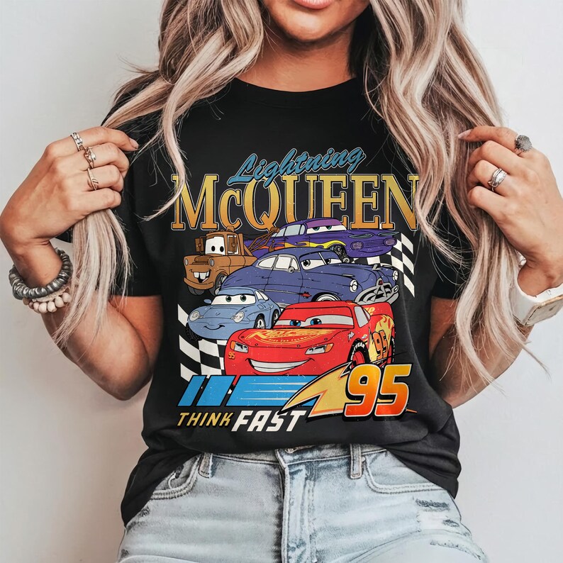 Puede incluir: Camiseta negra con un dise&ntilde;o de Lightning McQueen y otros personajes de Cars. El dise&ntilde;o incluye el texto "Lightning McQueen", "95" y "Think Fast". La imagen es colorida, con un fondo a cuadros.
