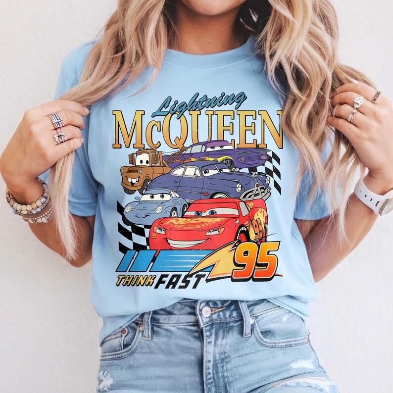 Puede incluir: Camiseta azul claro con un gr&aacute;fico de Rayo McQueen y otros personajes de Cars. El dise&ntilde;o incluye el texto "Lightning McQueen" y "Think Fast 95" en un estilo retro. La camiseta est&aacute; hecha de un material suave.
