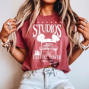May include: A maroon t-shirt featuring a white graphic of the Hollywood Studios Earffel Tower with Mickey Mouse ears. The text on the shirt reads "STUDIOS" above the tower and "EARFFEL TOWER CIRCA 1989 backlot tour" below.