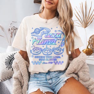 May include: Cream-colored t-shirt featuring a colorful graphic of the Pizza Planet logo, three alien characters, and the text "Serving Your Local Star Cluster Since 1995." The design has a retro, pastel aesthetic.