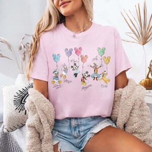 Pode incluir: Camiseta rosa claro com personagens da Disney segurando bal&otilde;es em forma de Mickey Mouse. Pato Donald, Margarida, Minnie, Mickey, Pateta e Pluto s&atilde;o retratados. Os nomes dos personagens est&atilde;o escritos abaixo.