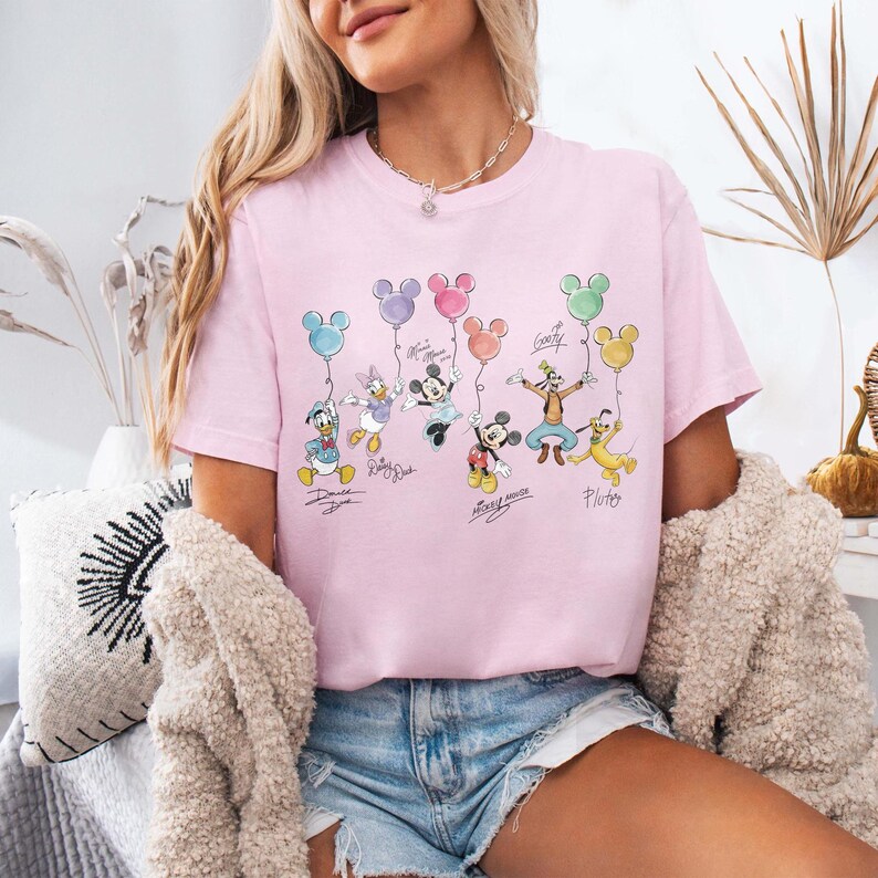 Pode incluir: Camiseta rosa claro com personagens da Disney segurando bal&otilde;es em forma de Mickey Mouse. Pato Donald, Margarida, Minnie, Mickey, Pateta e Pluto s&atilde;o retratados. Os nomes dos personagens est&atilde;o escritos abaixo.