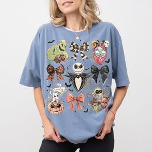 May include: A dusty blue short-sleeve t-shirt featuring a graphic print of characters and bows from The Nightmare Before Christmas, including Jack Skellington, Sally, Oogie Boogie, Zero, and Lock, Shock, and Barrel, arranged with small bats.
